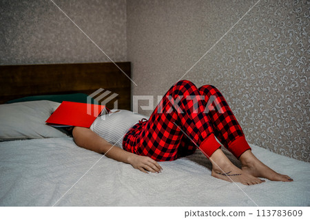 Balancing work, studies, and reading at home: Woman in red checkered pants lying on her bed with a red book and notes . Balancing work, studies, and reading at home: Woman in red checkered pants lying on her bed with a red book and notes . 113783609