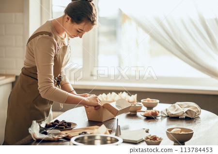 A chef in an apron works in a sunlit kitchen, folding and preparing a box filled with pastries and other delights. 113784453