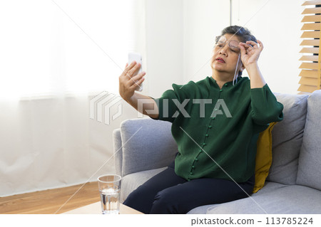 Elderly woman having troubles with long eye-sight trying to read a book, sitting on sofa at home. Elderly woman having troubles with long eye-sight trying to read a book, sitting on sofa at home. 113785224