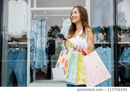 A woman holding shopping bags and a cellphone stands in front of a store window. She's a busy shopper using technology to communicate with friends and browse the latest trends. 113788299