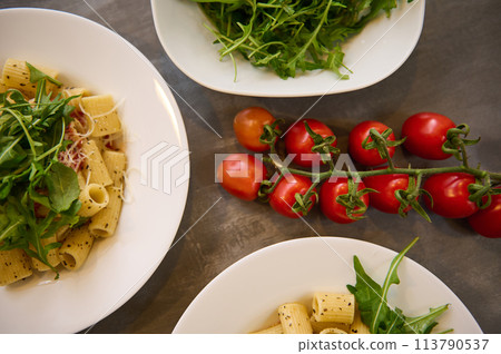 Pasta penne with tomato sauce, parmesan cheese nd arugula leaves. A branch of fresh ripe organic tomato cherry on the kitchen table. Top view 113790537