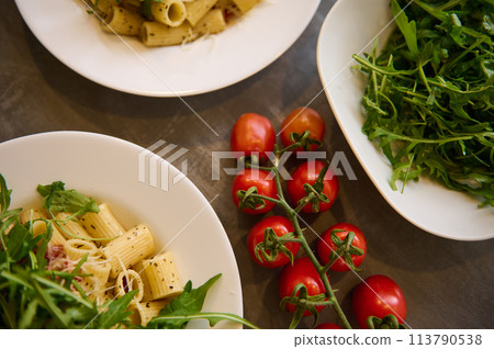 Food background with a branch of fresh ripe organic tomatoes cherry near plates of Italian pasta penne a la Amatriciana with pancetta bacon and pecorino cheese. Italian cuisine and culture. 113790538