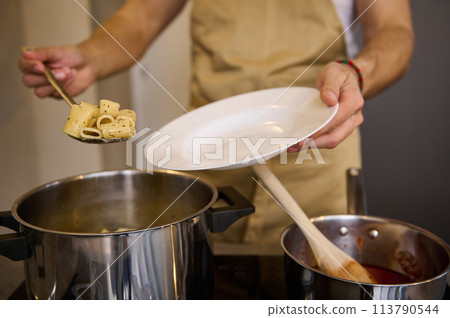 Close-up male chef standing at stove and putting freshly cooked pasta penne on a white plate 113790544