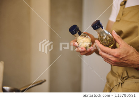 Close-up chef hands holding two glass bottles with condiments, dried basil leaves and ail for seasoning dish while cooking dinner 113790551