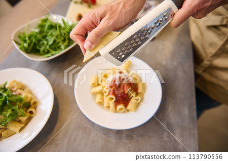 Directly above chef hands using grater, grating cheese on freshly cooked pasta with tomato sauce, seasoning and plating up the meal before serving it 113790556