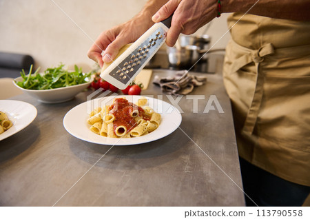 Close-up view of male chef plating up pasta before serving it to the customer. Hands holding grater, grating parmesan cheese, seasoning Italian pasta spaghetti while cooking dinner at home kitchen 113790558