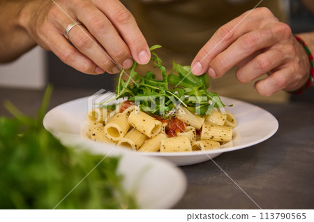 Close-up hands of a chef in home Kitchen, preparing and decorating meal with fresh arugula leaves, preparing pasta with tomato sauce according to traditional Italian recipe. Cuisine. Culinary. Epicure Close-up hands of a chef in home Kitchen, preparing and decorating meal with fresh arugula leaves, preparing pasta with tomato sauce according to traditional Italian recipe. Cuisine. Culinary. Epicure 113790565