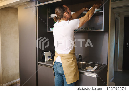 Young man in a chef's apron in a home kitchen taking out plates from the kitchen cabinet 113790674