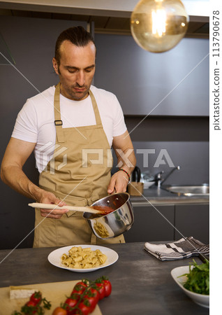 Vertical shot of attractive European male chef pouring tomato sauce on Italian pasta, plating up the dish before serving. Man cooking dinner at home kitchen. Cuisine. Culinary. Epicure. Food concept 113790678