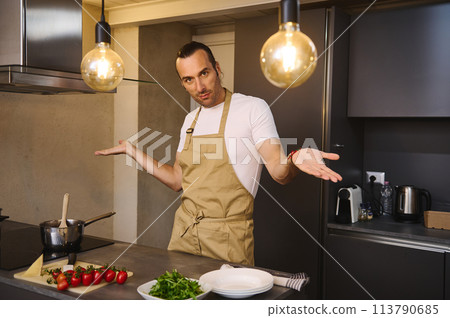 Handsome Caucasian man in chef's apron cooking family dinner, smiling looking at camera Handsome Caucasian man in chef's apron cooking family dinner, smiling looking at camera 113790685