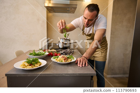 Attractive European male chef decorating spaghetti Bolognese with fresh greens, standing at kitchen island in the home kitchen. Man cooking dinner at home 113790693