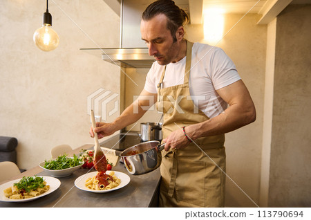 European professional chef plating up pasta before serving it to the customer. Handsome man in apron, holding stainless steel pan and pouring tomato sauce on pasta, cooking dinner at home kitchen 113790694