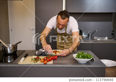 European man in beige chef's apron, standing at kitchen counter and using kitchen towel, removing traces of tomato sauce on a plate of freshly prepared Italian pasta. People. Culinary. Italian cuisine 113790698