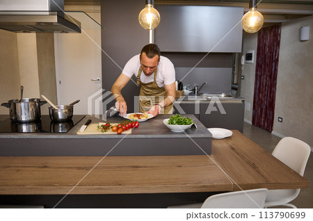 Portrait of a man in chef's apron, standing at kitchen island and using kitchen towel, wiping away traces of tomato sauce on a plate of freshly prepared spaghetti. People. Culinary. Italian cuisine Portrait of a man in chef's apron, standing at kitchen island and using kitchen towel, wiping away traces of tomato sauce on a plate of freshly prepared spaghetti. People. Culinary. Italian cuisine 113790699