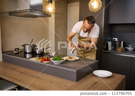 Male chef cooking in the home kitchen, grates parmesan cheese over a plate with freshly boiled pasta with tomato sauce. Ingredients on the table in stylish modern minimalist home kitchen interior Male chef cooking in the home kitchen, grates parmesan cheese over a plate with freshly boiled pasta with tomato sauce. Ingredients on the table in stylish modern minimalist home kitchen interior 113790705