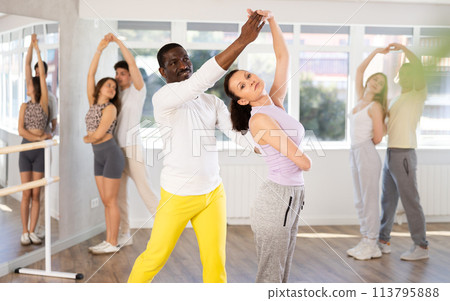 Smiling young man and woman with multinational group active people training energetic active swing dance movements in dance salon 113795888
