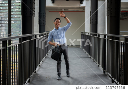 A friendly Asian businessman waving to greet his colleague while walking along the building corridor A friendly Asian businessman waving to greet his colleague while walking along the building corridor 113797636