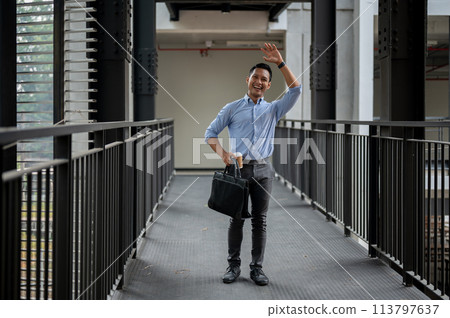 A friendly Asian businessman waving to greet his colleague while walking along the building corridor A friendly Asian businessman waving to greet his colleague while walking along the building corridor 113797637