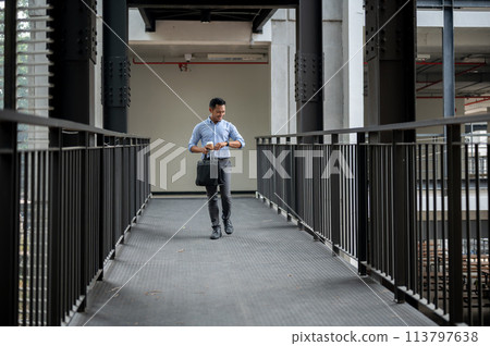 A confident Asian businessman is checking time on his wristwatch while walking along a corridor. 113797638