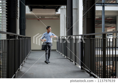 A confident Asian businessman is checking time on his wristwatch while walking along a corridor. A confident Asian businessman is checking time on his wristwatch while walking along a corridor. 113797639