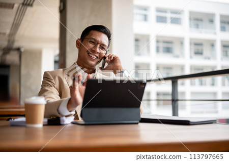 A confident businessman sits at a wooden table with a tablet and documents, talking on the phone. A confident businessman sits at a wooden table with a tablet and documents, talking on the phone. 113797665
