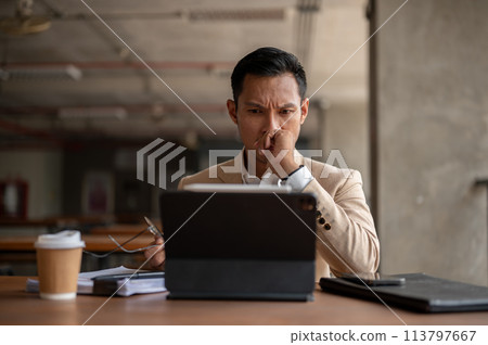 An Asian businessman is staring at his tablet screen with a serious expression, facing a problem. 113797667