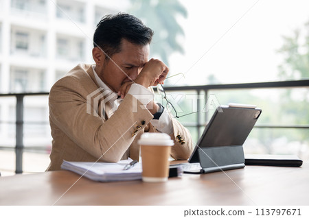 A frustrated Asian businessman covers his face with his hands while looking at a digital tablet. A frustrated Asian businessman covers his face with his hands while looking at a digital tablet. 113797671