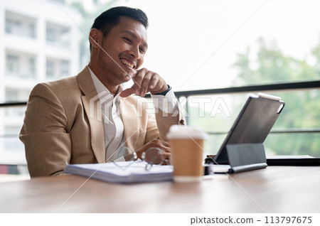 A confident Asian businessman sits at a table, working on his digital tablet and document. A confident Asian businessman sits at a table, working on his digital tablet and document. 113797675