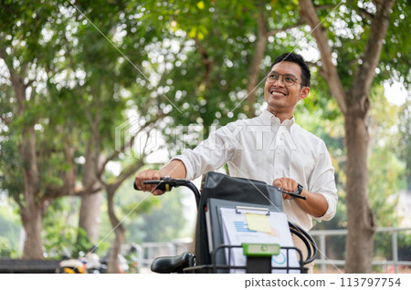 A happy, carefree Asian businessman pushing his bike, walking in the public park. A happy, carefree Asian businessman pushing his bike, walking in the public park. 113797754