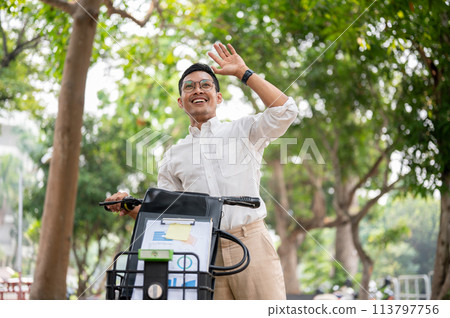 A happy businessman smiles and waves while pushing his bike, greeting someone while heading to work. A happy businessman smiles and waves while pushing his bike, greeting someone while heading to work. 113797756