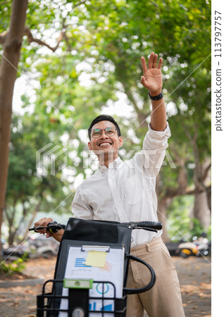 A happy businessman smiles and waves while pushing his bike, greeting someone while heading to work. A happy businessman smiles and waves while pushing his bike, greeting someone while heading to work. 113797757