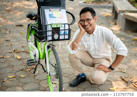 A happy, smiling Asian businessman sits next to his bicycle in a park, going to work in the morning. A happy, smiling Asian businessman sits next to his bicycle in a park, going to work in the morning. 113797758