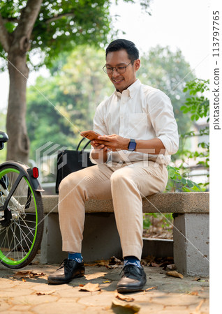 A happy Asian businessman sits on a bench in a public park, using his smartphone. A happy Asian businessman sits on a bench in a public park, using his smartphone. 113797765