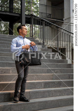 An exhausted Asian millennial businessman stands on the stairs, holding a coffee cup and a briefcase An exhausted Asian millennial businessman stands on the stairs, holding a coffee cup and a briefcase 113797790