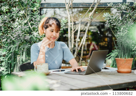 Contemplative woman works on her laptop at a wooden garden table, a serene green setting with plants and sunlight enhancing the calm work environment. 113800682