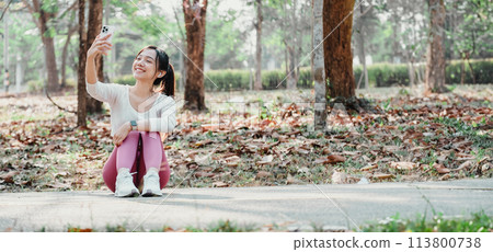 Seated woman in pink leggings takes a selfie with her smartphone in a park filled with fallen leaves and trees. Seated woman in pink leggings takes a selfie with her smartphone in a park filled with fallen leaves and trees. 113800738