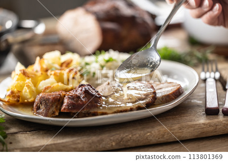 A close-up view of a spoon with sauce that pours over the meat, roasted neck. As a side dish to the meat are baked potatoes and jasmine rice. 113801369