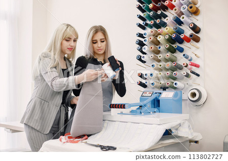 Two dressmakers standing in workshop near the bright wall with threads Two dressmakers standing in workshop near the bright wall with threads 113802727