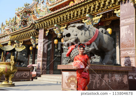 Portrait pretty Asian female in red traditional Chinese costume holding a fan standing and posing at stone lion  in the shrine. 113804678