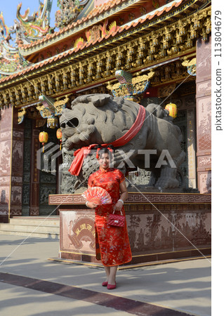 Portrait pretty Asian female in red traditional Chinese costume holding a fan standing and posing at stone lion  in the shrine. 113804679