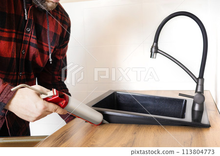 A man installs a black stone sink into a kitchen countertop. Worker seals up the kitchen sink with a sealant using a construction sealing gun A man installs a black stone sink into a kitchen countertop. Worker seals up the kitchen sink with a sealant using a construction sealing gun 113804773