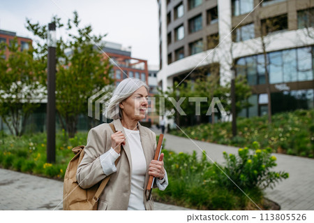 Mature businesswoman scrolling on smartphone, going home from work. Beautiful older woman with gray hair walking down city street. 113805526
