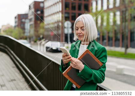 Mature businesswoman scrolling on smartphone, going home from work. Beautiful older woman with gray hair standing on city street. Mature businesswoman scrolling on smartphone, going home from work. Beautiful older woman with gray hair standing on city street. 113805532