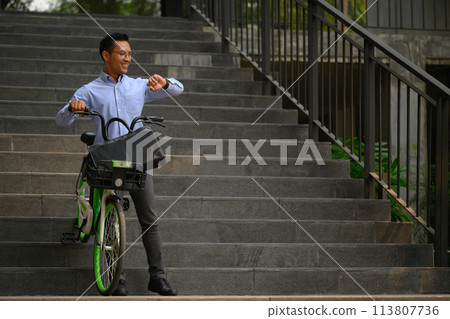 Smiling businessman with his bicycle in front of the building and checking time on wristwatch 113807736