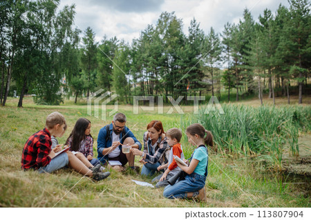 Teacher showing sample of lake water to school children, during field teaching class. Outdoor active education helping young student to learn about ecosystem. Teacher showing sample of lake water to school children, during field teaching class. Outdoor active education helping young student to learn about ecosystem. 113807904