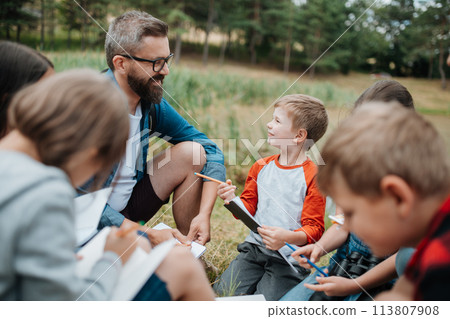 Young students learning about nature, forest ecosystem during biology field teaching class, writing notes. Teachers talking with children during outdoor active education. 113807908
