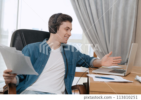 Cheerful young guy in headphones having video conferevce at home Cheerful young guy in headphones having video conferevce at home 113810251
