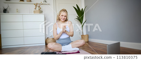 Portrait of young charismatic fitness trainer, girl blogger records video on digital camera, talks about health and workout, doing exercises on rubber mat in a room at home 113810375