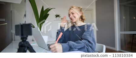 Portrait of young blond smiling woman, studying at home, remote education concept, connects to online course or lesson, writing in notebook 113810384