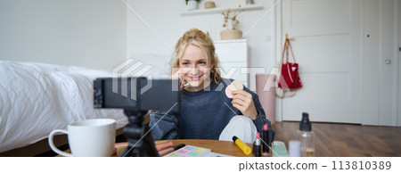 Image of young woman, vlogger records video on digital camera, shows beauty products, recommending makeup for audience on social media, sits on front in a room Image of young woman, vlogger records video on digital camera, shows beauty products, recommending makeup for audience on social media, sits on front in a room 113810389